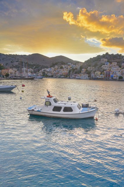 Fishing boat moored in tranquil harbor at sunset with scenic coastal town and mountains in background. Ideal for travel blogs, tourism promotions, and posters depicting serene vacation destinations.