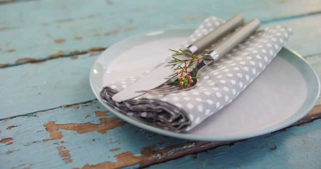 A neatly arranged table setting features a white plate with polka dots, silver cutlery wrapped in a gray napkin, adorned with a sprig of greenery, on a rustic blue wooden table, with copy space. The setting evokes a casual yet elegant dining atmosphere, ideal for a festive or special occasion.