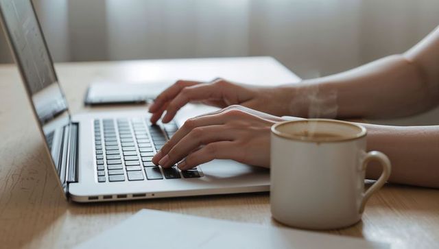 Typing on laptop at home desk with steaming coffee cup by soft curtain light work routine