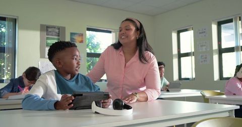 Teacher assisting african american boy using tablet for learning in bright classroom