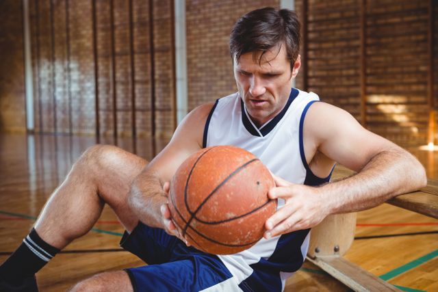 Basketball player sitting on court holding ball, reflecting. Ideal for sports motivation, athletic training, and basketball-related content.