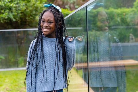 Smiling african american preteen girl leaning on glass railing, garden reflection and candid joy