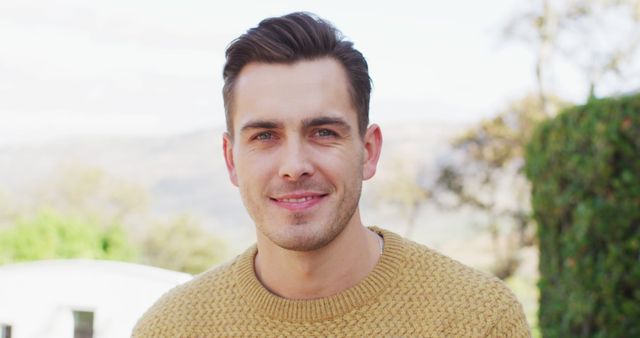 Portrait of a smiling man with short dark hair wearing a yellow sweater standing outdoors. This can be used for portraying confidence, casual fashion, positivity, lifestyle blogs, and advertisements focusing on outdoor activities or happiness.