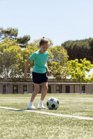 Young girl practicing soccer dribbling on sunny turf field with trees background