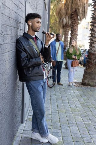Young man leaning on brick wall speaking into smartphone with earbuds on urban sidewalk