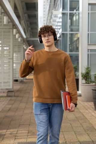 Young student walking on modern campus walkway wearing headphones and holding notebooks