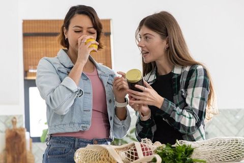 Diverse female friends unpacking groceries in kitchen, sniffing jar and reading label
