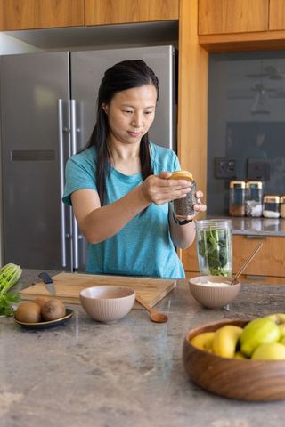 Asian woman preparing green smoothie in modern kitchen holding seed jar with fresh ingredients