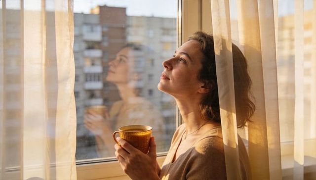 Woman gazing through window holding warm mug in golden morning light, cozy apartment