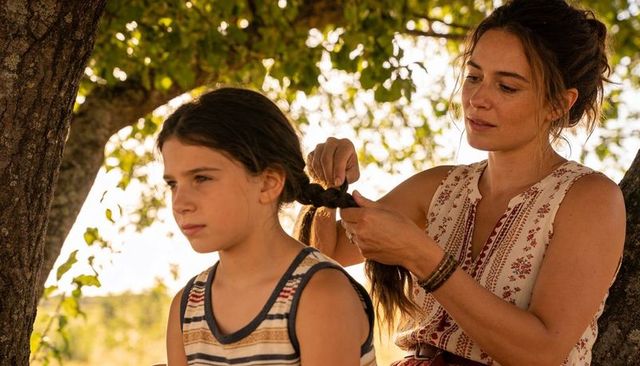 Mother braiding daughter hair under sunlit tree during golden hour family bonding moment