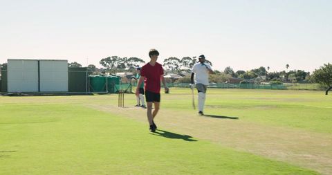 Cricket bat resting on suburban grass pitch near stumps with players walking in sunshine