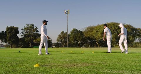Cricket team practicing bowling and fielding on grassy pitch with training cones and coach