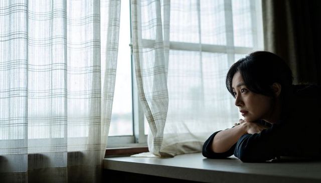 Leaning asian woman gazing through sheer plaid curtains, contemplating quiet apartment