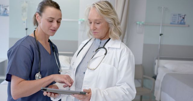 Female medical professionals are standing in a hospital room, engaged in a discussion while using a digital tablet. Both appear to be reviewing patient information or medical data. The background features medical equipment and a clinical setting. Suitable for use in healthcare publications, medical technology articles, or promotional materials related to hospitals and medical services.