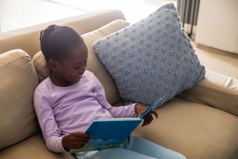 African american girl reading picture book on beige sofa, smiling and relaxing in sunlight