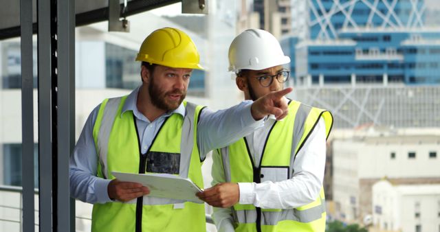 Two male engineers wearing safety vests and hard hats are actively discussing a construction project on site. One is holding blueprints and both are engaged in conversation, pointing towards a particular direction. This image is ideal for illustrating teamwork, professional engineering, and construction management in corporate presentations, industry reports, or safety training materials.