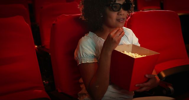 Young girl watches a 3D movie at a cinema, wearing 3D glasses and eating popcorn. Surroundings include red theater seats, highlighting sense of immersive experience and enjoyment. Ideal for illustrating family entertainment, movie theaters, children at the movies, or leisure activities.