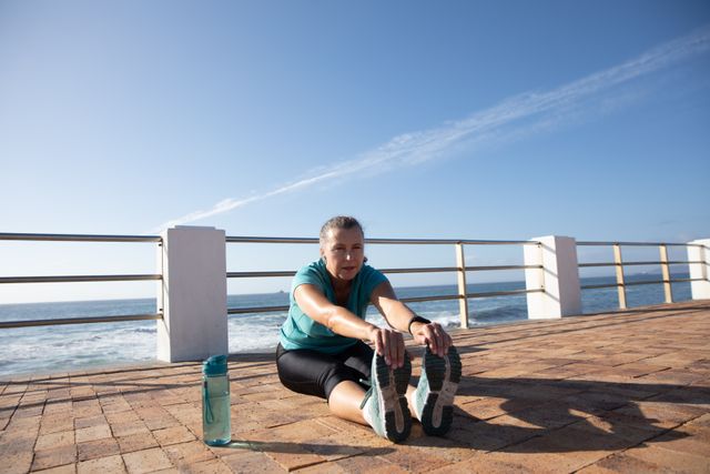 Senior woman stretching her legs on a seaside promenade, wearing sports clothes and with a water bottle next to her. Ideal for promoting healthy lifestyles, retirement activities, fitness routines, and wellness programs for seniors. Perfect for use in health and fitness blogs, retirement community advertisements, and wellness campaigns.