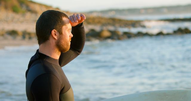 A man in a wetsuit gazes at the ocean while holding a surfboard. The setting includes a rocky coastline and gentle waves under a clear blue sky. Perfect for promoting outdoor activities, surfing tutorials, beachwear advertisements, or adventure travel blogs.