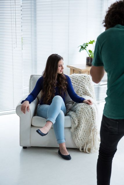 Female model sitting on a white chair in a studio, posing for a photoshoot. She is wearing a casual outfit with a blue top and light jeans. The setting includes natural light, a cozy blanket, and a modern interior. This image can be used for fashion magazines, lifestyle blogs, or promotional materials for photography services.
