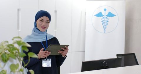 African american healthcare receptionist wearing hijab holding tablet and smiling at clinic desk