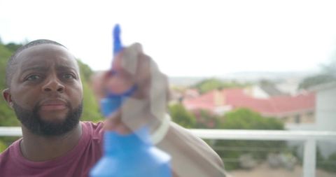 African man cleaning balcony glass with blue spray and cloth, outdoor terrace closeup