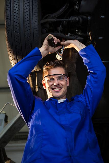 Mechanic performing maintenance on car undercarriage, wearing blue uniform and safety glasses, working with tools. Ideal for illustrating automotive services, repair industry, professional maintenance work, and mechanic expertise.