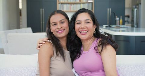 Two women smiling and embracing on sofa in modern kitchen, cozy lifestyle portrait