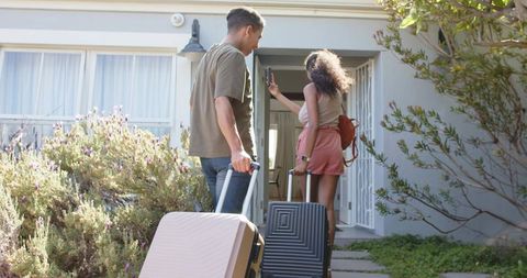African american couple arriving at vacation rental carrying luggage and using phone