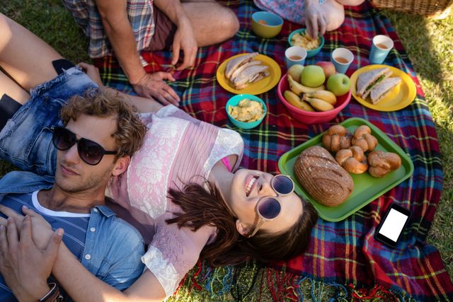 Group of friends enjoying a relaxing picnic in the park on a sunny day. They are lying on a colorful blanket with a spread of food including sandwiches, fruit, and pastries. Perfect for use in content related to outdoor activities, leisure time, summer gatherings, friendship and bonding moments.