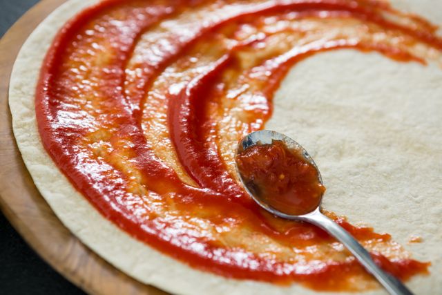 Close-up view of pizza dough with tomato sauce being spread using a spoon. Ideal for use in culinary blogs, recipe websites, cooking tutorials, and food-related advertisements. Perfect for illustrating the process of making homemade pizza or showcasing Italian cuisine.