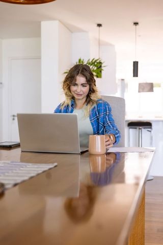 Woman Enjoying Remote Work with Laptop and Coffee in Bright Home Kitchen