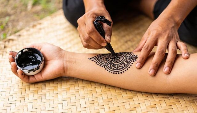 Henna artist applying intricate mandala on forearm with paste cone and jar on woven mat