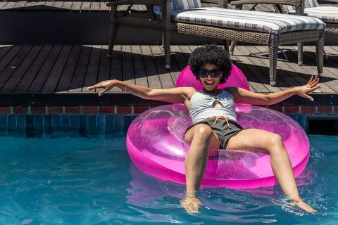Smiling woman floating on pink inflatable ring relaxing at sunny backyard pool deck