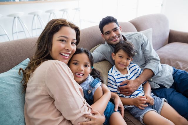 This image shows a happy family of four sitting on a sofa in their living room. The parents and children are smiling and appear to be enjoying their time together. This image can be used for family-oriented advertisements, parenting blogs, home decor promotions, or articles about family life and bonding.