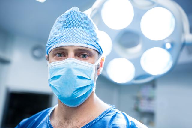 Surgeon wearing a surgical mask and scrubs in an operating room, illuminated by surgical lights. Ideal for use in healthcare-related content, medical articles, hospital brochures, and educational materials about surgery and medical professions.
