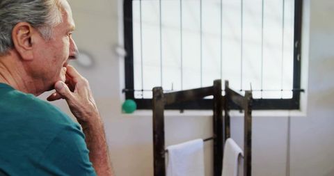 Senior man touching chin while contemplating by bathroom towel rack near barred window