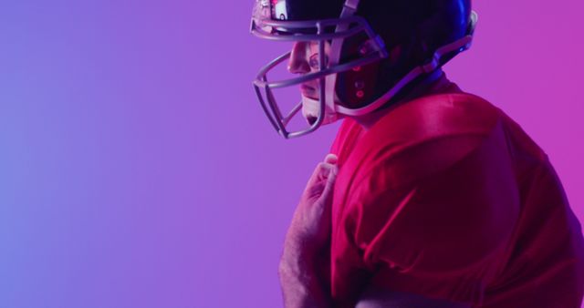 Football player fully geared and holding his chest, captured under vibrant, colorful studio lights. This can be used for sports promotions, athletic equipment advertisements, or campaign posters emphasizing strength and determination in football. The contrast of the colorful lighting highlights intense emotions and draws focus to the action.