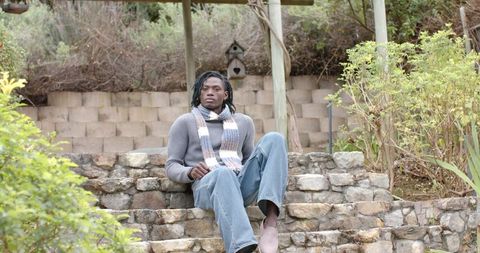 Young man sitting on stone garden steps wearing cozy sweater and striped scarf