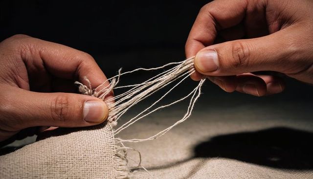 Hands pulling loose threads from frayed linen edge for textile repair