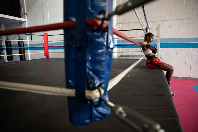 Biracial female boxer practicing in a boxing gym wearing sports clothes, resting on boxing ring. Strength sports achievement.