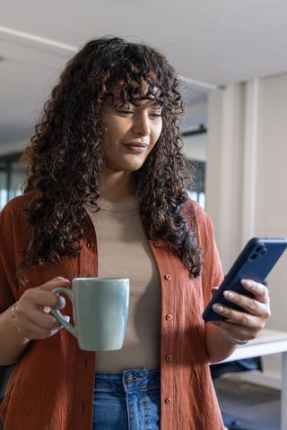 Young professional woman holding mug and checking smartphone in modern office environment
