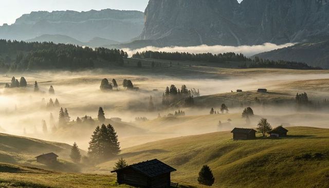 Morning mist rolling over alpine meadow with rustic cabins and mountain massif