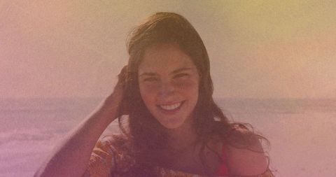 Smiling young woman touching hair at sunlit beach, pastel grain and coastal haze, carefree