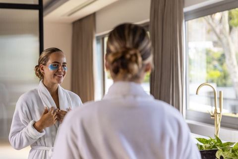 Woman wearing blue under-eye gel patches in white robe looking in mirror morning skincare