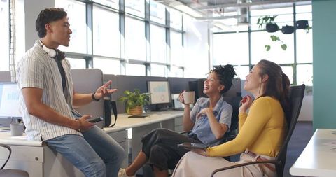 Diverse coworkers chatting and laughing in modern open-plan office with devices and coffee