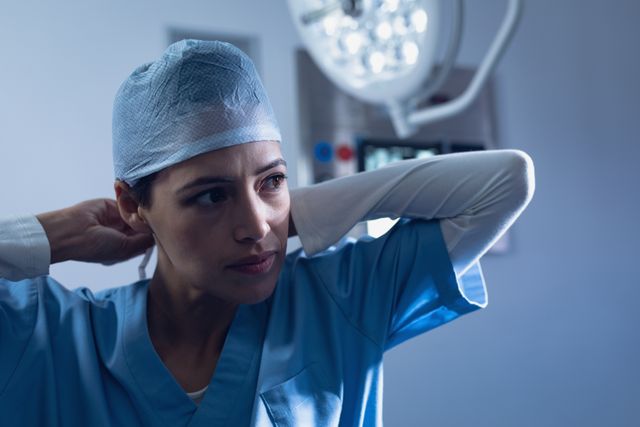 Image shows a female surgeon in blue scrubs adjusting her surgical cap in a hospital's operation room. Ideal for use in healthcare-related articles, hospital brochures, medical websites, or educational materials highlighting the roles and responsibilities of medical professionals.