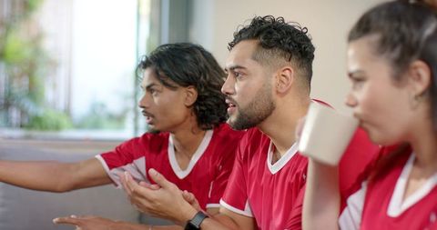 Friends watching live soccer match wearing red jerseys on living room couch, tense moment