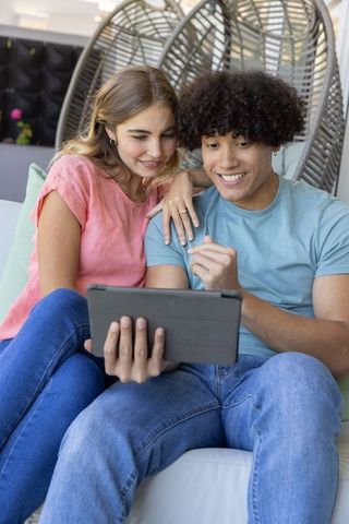 Young couple smiling while browsing tablet on sofa, woman showing ringed hand