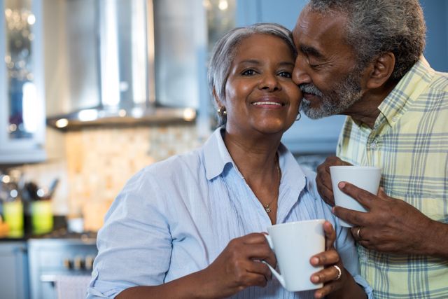 Senior couple sharing a tender moment in their kitchen while holding coffee mugs. Ideal for use in advertisements promoting senior living, retirement communities, healthcare services, or lifestyle blogs focusing on relationships and family life.